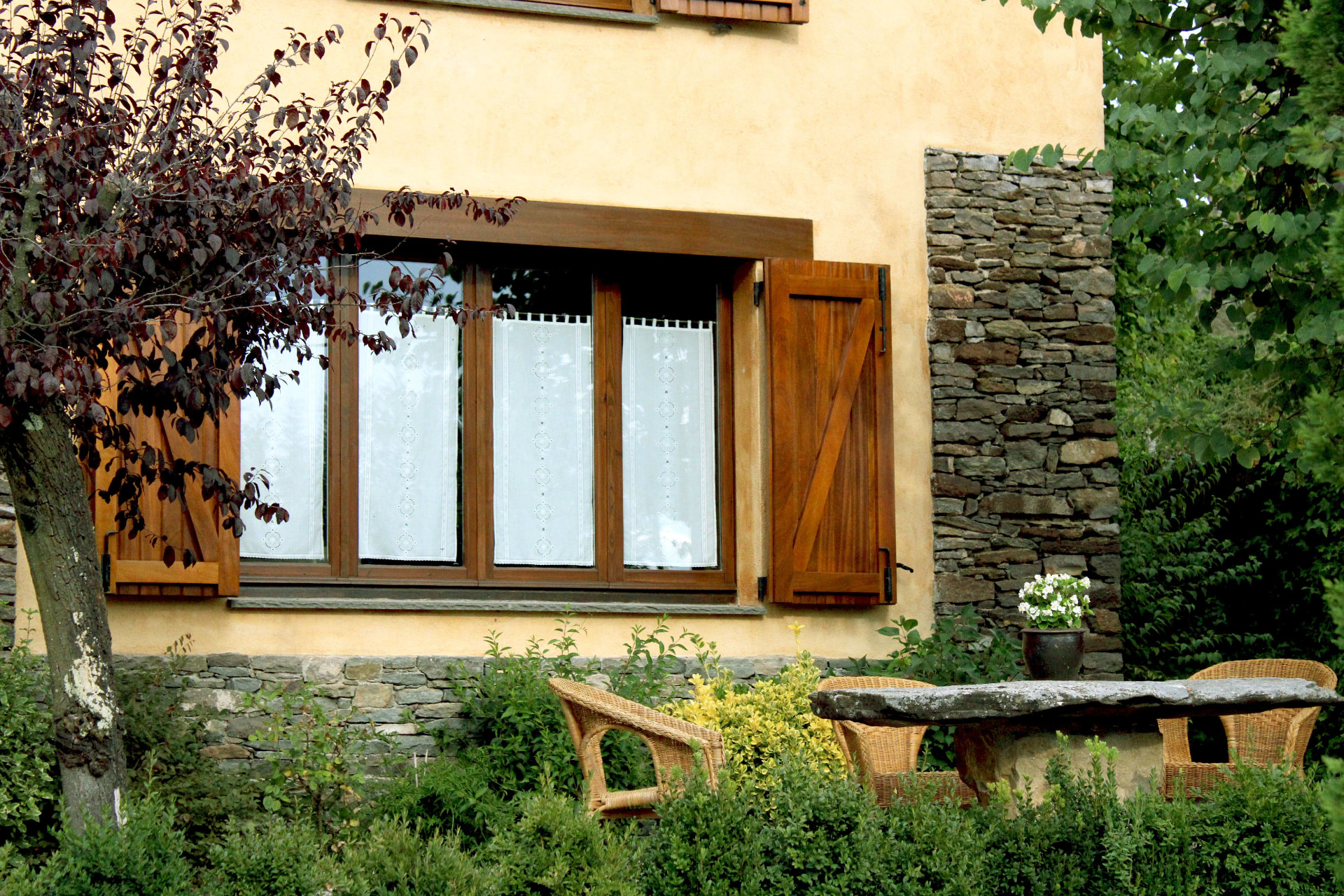 Finestra con imposte in legno scuro su una facciata ocra, affaccio su un giardino con piante e mobili da esterno