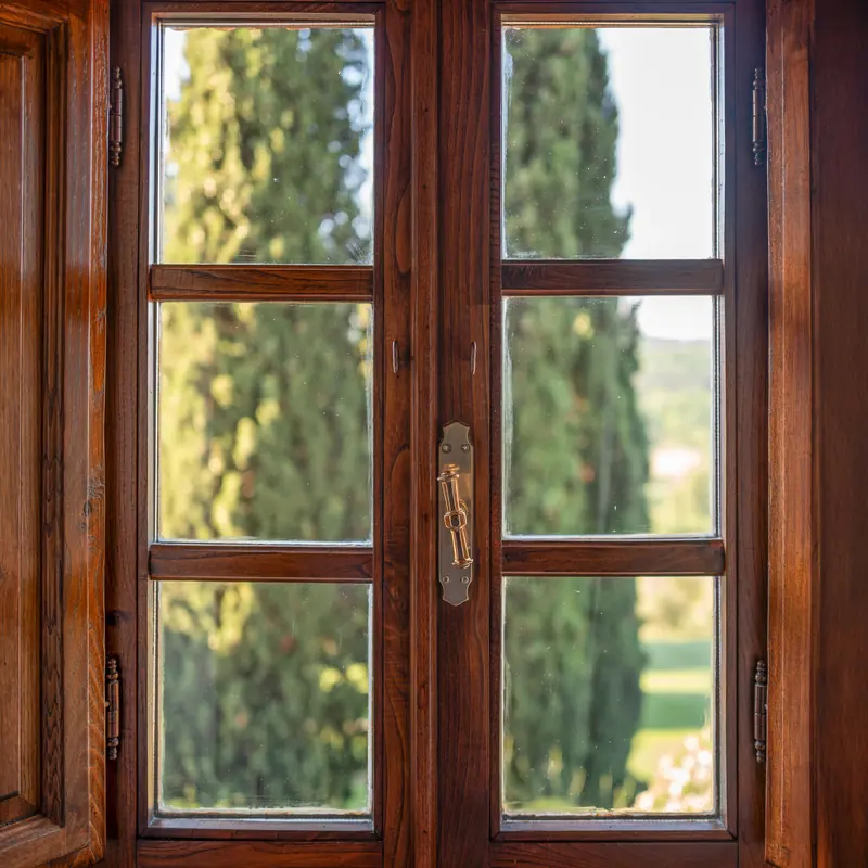 Finestra in legno con maniglia in ottone affacciata su cipressi verdeggianti