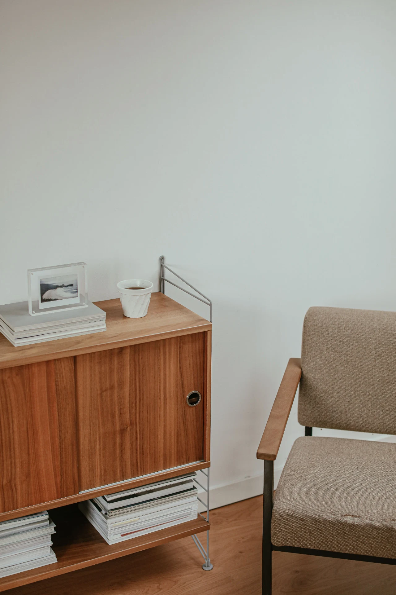 Angolo di casa con mobiletto in legno, libri e tazza, e poltroncina con braccioli in legno